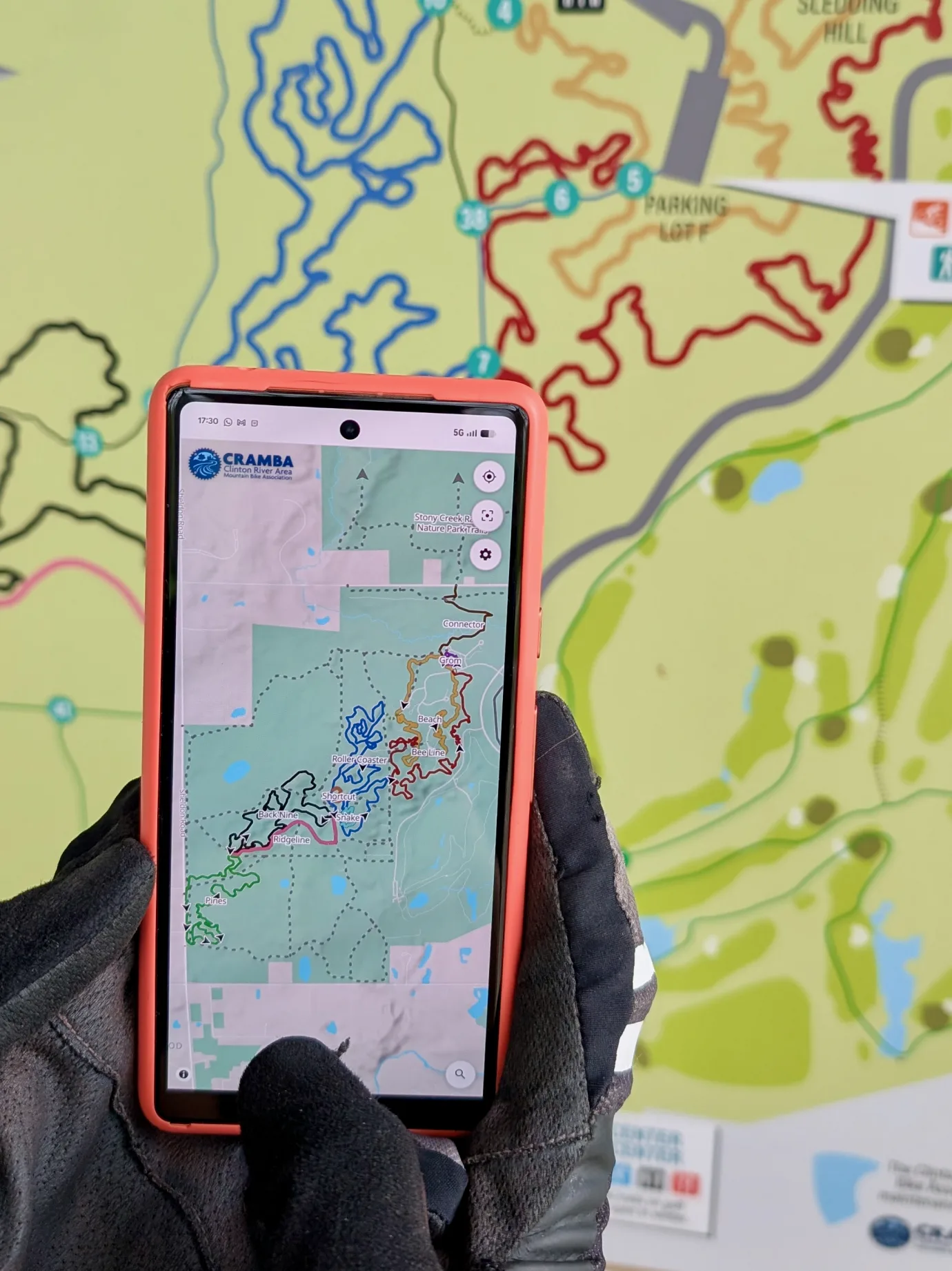 A phone running trailmaps.app, held in front of a trailhead's printed map board, with the on-screen trails matching the colors on the sign